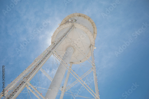 Looking up at a bright white water tower from below backlit with the sun and a bright blue sky.