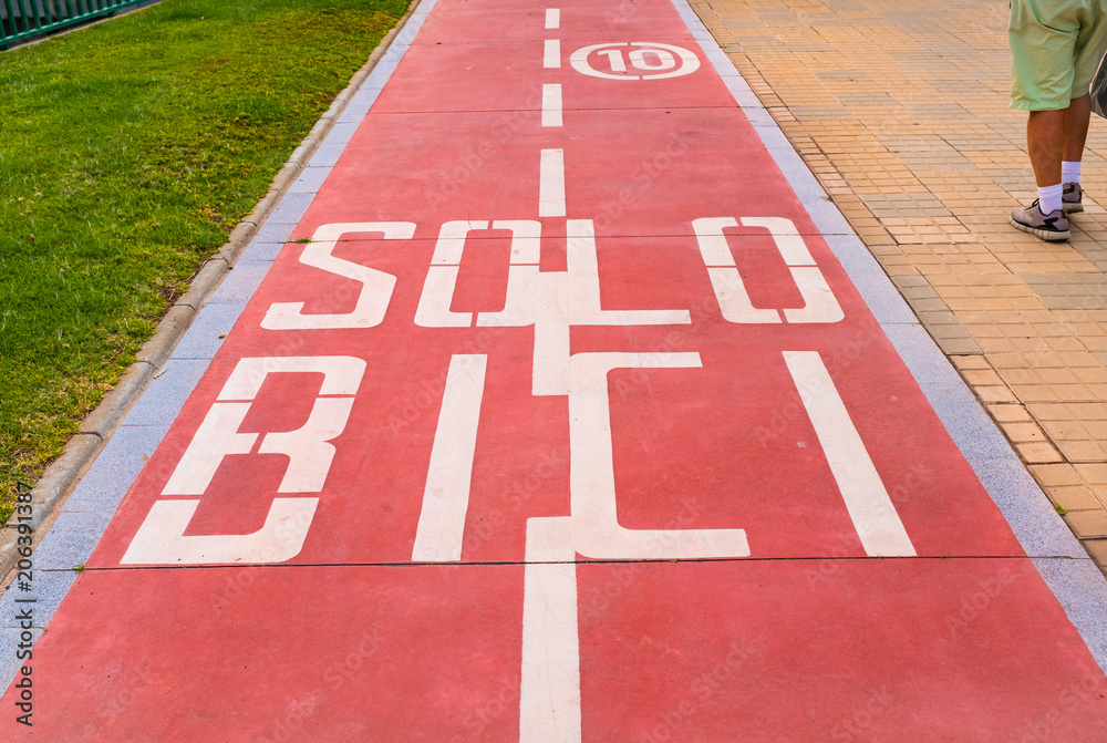 Red bicycle lane in Spain. Saying Solo Bici, Only Bikes at a max speed of 10 Kmph. There is a man walking in shorts next to the path
