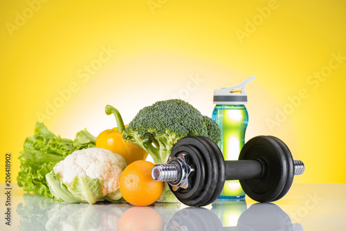 dumbbell, bottle of water and fresh orange and vegetables on yellow