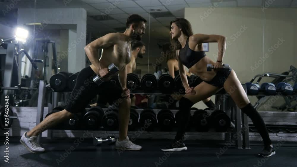 The man and the woman of sports bodywork are engaged in a sports hall doing exercises for hands with dumbbells, two trainings in a sports hall work out exercises on hands a side view, dumbbells for The man and the woman of sports bodywork are engaged in a sports hall doing exercises for hands with dumbbells, two trainings in a sports hall work out exercises on hands a side view, dumbbells for