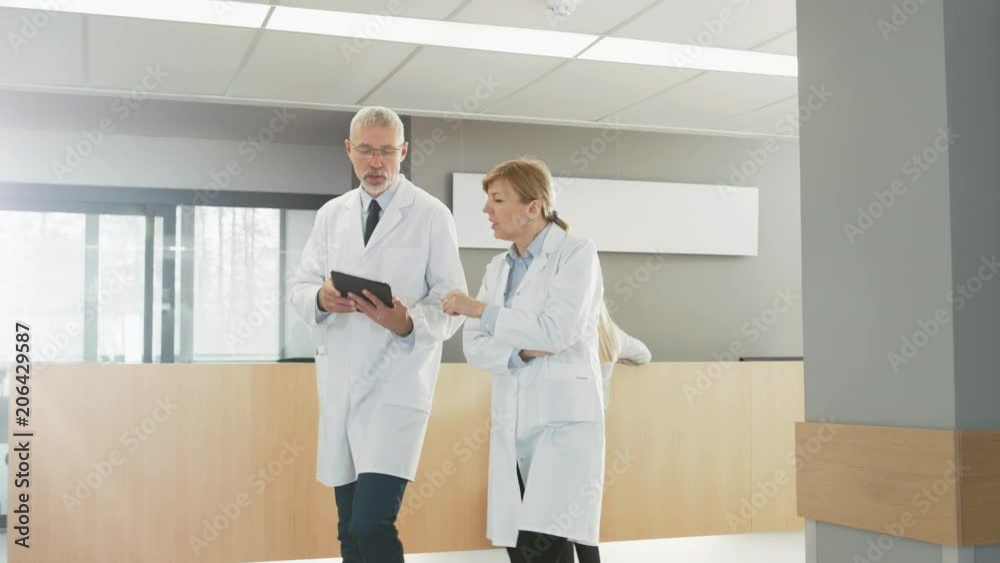 In the Hospital, Busy Doctors Talk, Using Tablet Computer While Walking Through the Building. In the Background Patient Talks with Receptionist.  Shot on RED EPIC-W 8K Helium Cinema Camera.