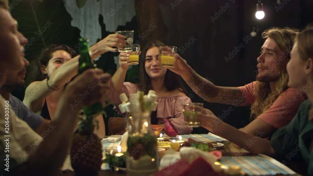Group of young friends sitting at table with tropical fruits on it, talking to each other and clinking drinks