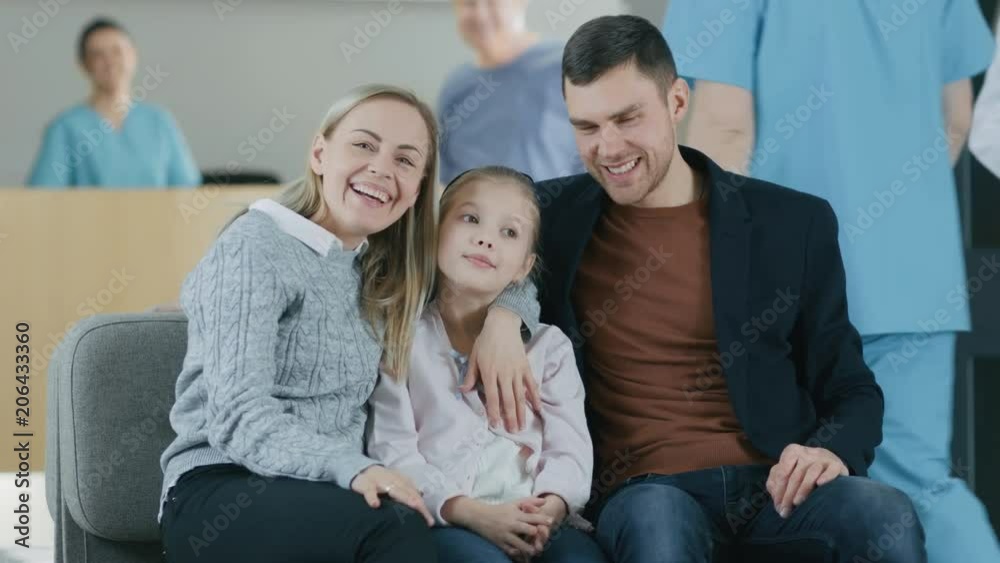 Portrait of the Young Happy Family in the Hospital. Handsome Father, Beautiful Mother and Cute Little Daughter Sitting in the Lobby of the Medical Facility, Smiling. Portrait of the Young Happy Family in the Hospital. Handsome Father, Beautiful Mother and Cute Little Daughter Sitting in the Lobby of the Medical Facility, Smiling.