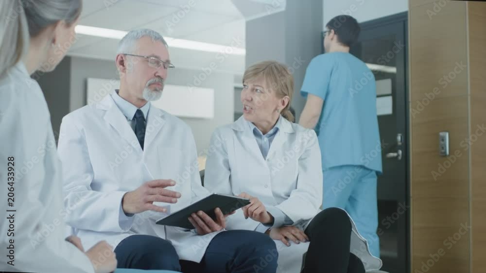 In the Lobby of the Hospital Three Professional Doctor Have Discussion while Sitting. They Use Tablet Computer. Slow Motion. Shot on RED EPIC-W 8K Helium Cinema Camera. In the Lobby of the Hospital Three Professional Doctor Have Discussion while Sitting. They Use Tablet Computer. Slow Motion. Shot on RED EPIC-W 8K Helium Cinema Camera.