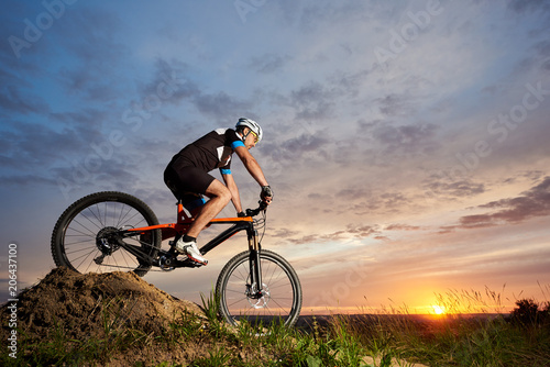 View of energetic and active man in helmet and sportswear riding orange bike in the evening. Sporty and energetic athlete sitting and rolling down hill at sunset. Recreative fresh air activities. © anatoliy_gleb