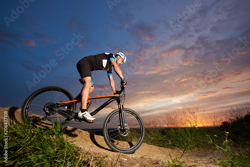 Guy wearing a helmet and sportswear on a mountain bike coming down the hill under the evening sky against the sun at sunset. The concept of an active lifestyle