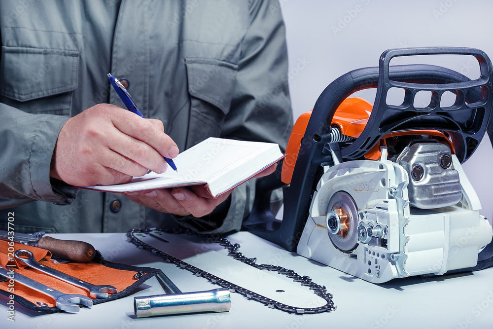 Professional technician working in repair service. Repairing chainsaw on workbench.Repairman records work plan in notebook. Professional technician working in repair service. Repairing chainsaw on workbench.Repairman records work plan in notebook.
