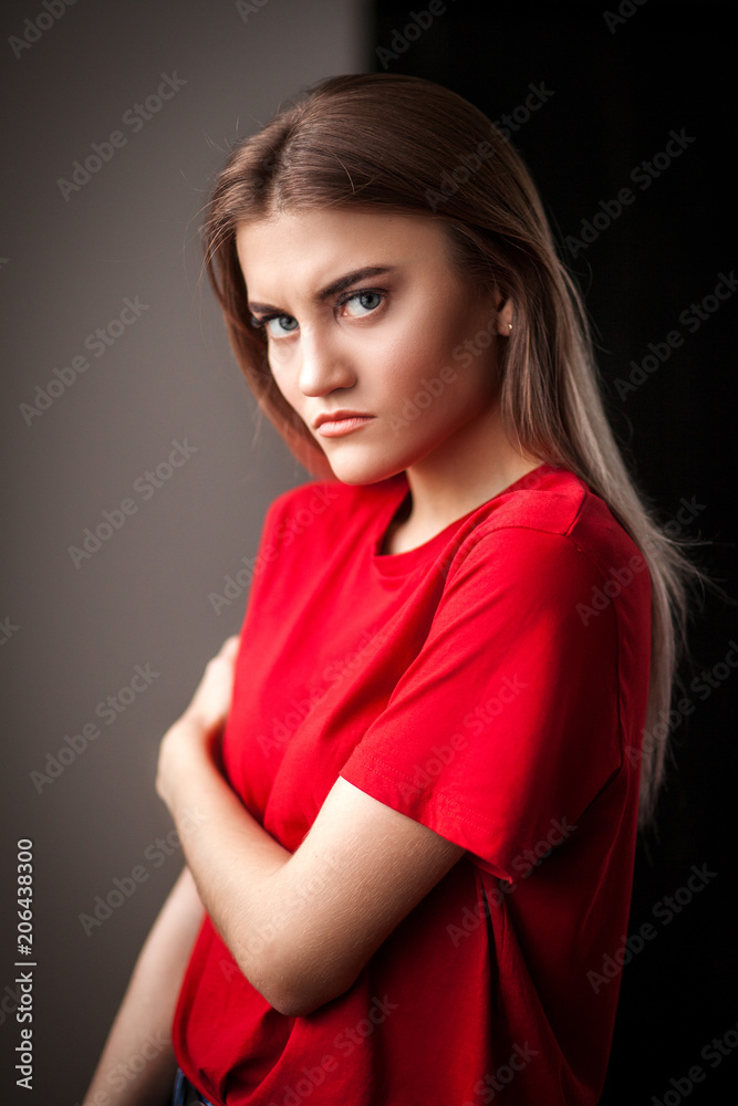 Portrait serious beautiful young woman in red t-shirt and jeans posing with hands on hip and looking at camera in studio on black background Portrait serious beautiful young woman in red t-shirt and jeans posing with hands on hip and looking at camera in studio on black background