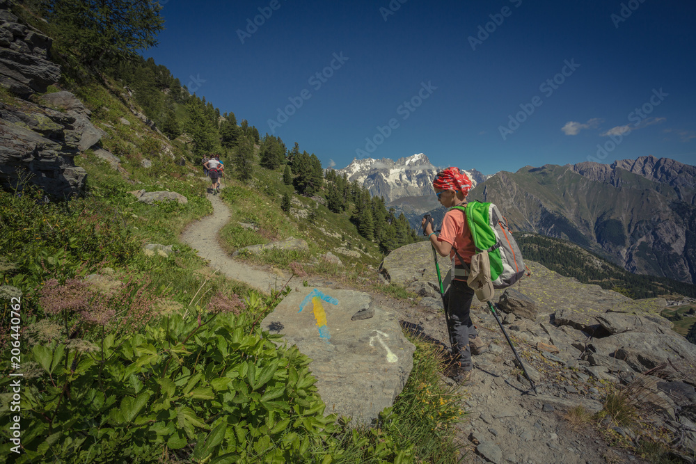 A young boy hiking on a mountain path on Italian alps. A male with hat and backpack, holds trekking poles in her hands. Relaxing alone in a beautiful summer sunny day. A kid enjoying outdoor sport.