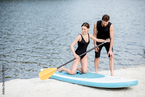 Male coach teaching young woman to surf on the standup paddleboard with oar standing on the beach