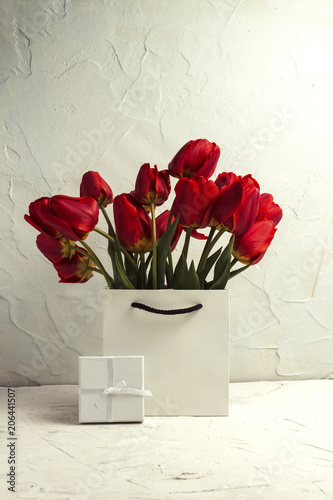 Fototapeta Naklejka Na Ścianę i Meble -  White Gift Bag, small white gift box, petals and bouquet of red tulips on a light stone background. Concept Offer a gift or an engagement, marriage