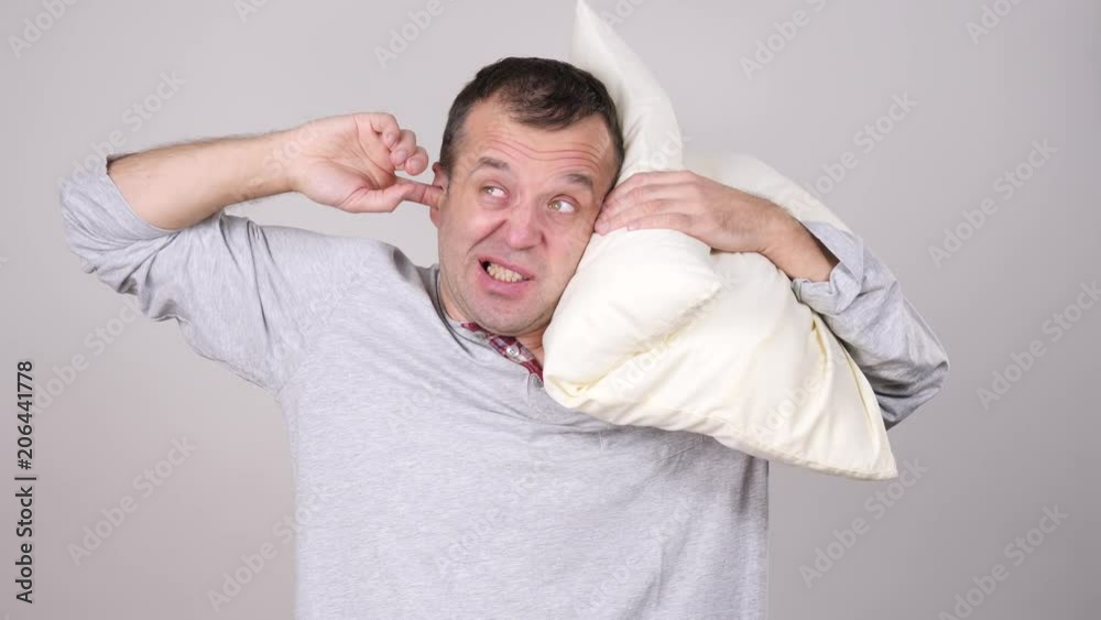 Sleepy tired angry guy holding pillow in one and closing his ear with another hand protecting from noise, on grey. Health care, sleep disorder concept. Sleepy tired angry guy holding pillow in one and closing his ear with another hand protecting from noise, on grey. Health care, sleep disorder concept.