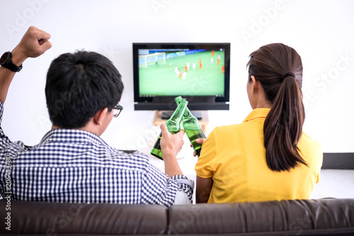 Young Asian couple love watching soccer match on tv and cheering football team, celebrating with beer and popcorn at home, sports and entertainment concept