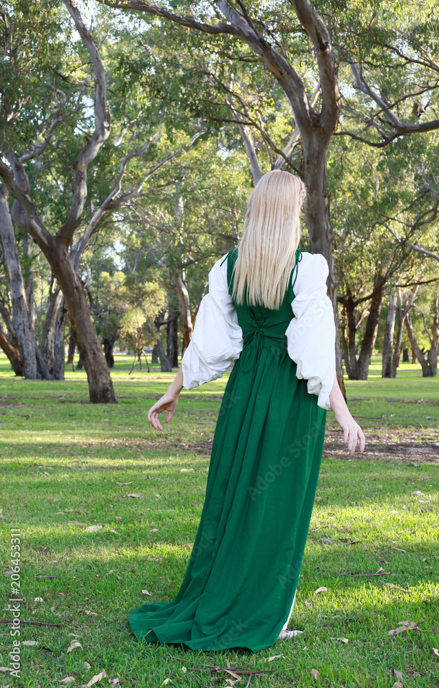 full length portrait of blonde girl wearing greensand white medieval costume, standing pose. walking through a forest.