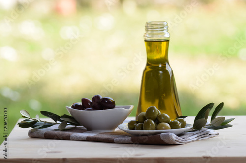 bottle of oil and two bowls of black and green olive on a table outside