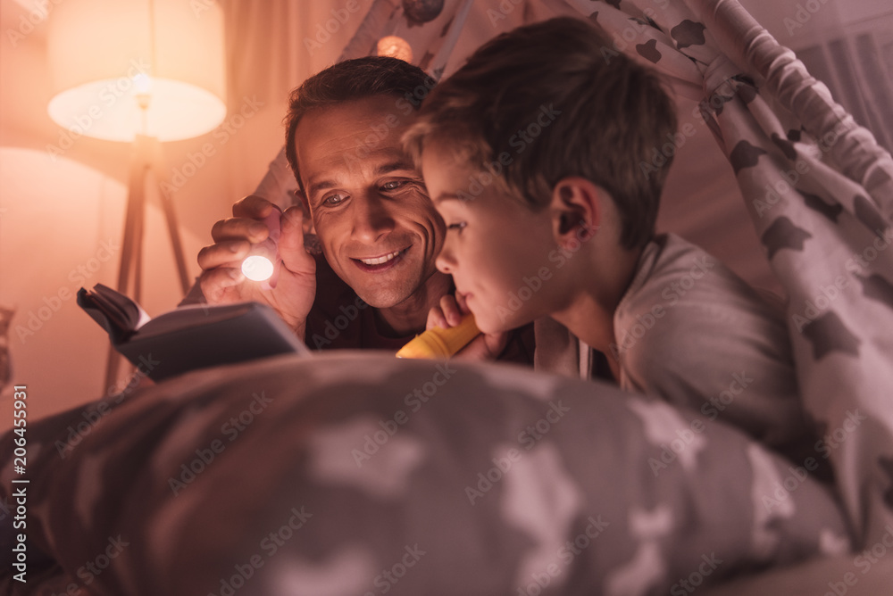 Before sleep. Nice pleasant caring father holding a flash light and smiling  while reading a fairy tale to his son Stock Photo | Adobe Stock