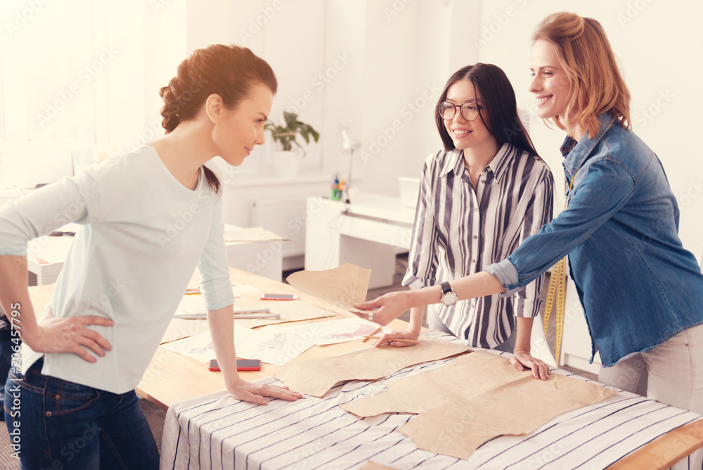 Choice yours. Young pleasant dressmaker giving clothing shape to her colleague while the colleague leaning on the table and expressing interest