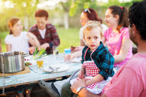 Little boy with adorable little apron sitting on his father lap and looking at the distance while the rest of the family is sitting eating at the picnic table and eating.