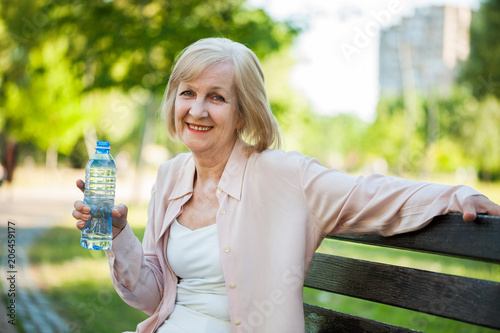 Adult woman is sitting in park and drinking water. 