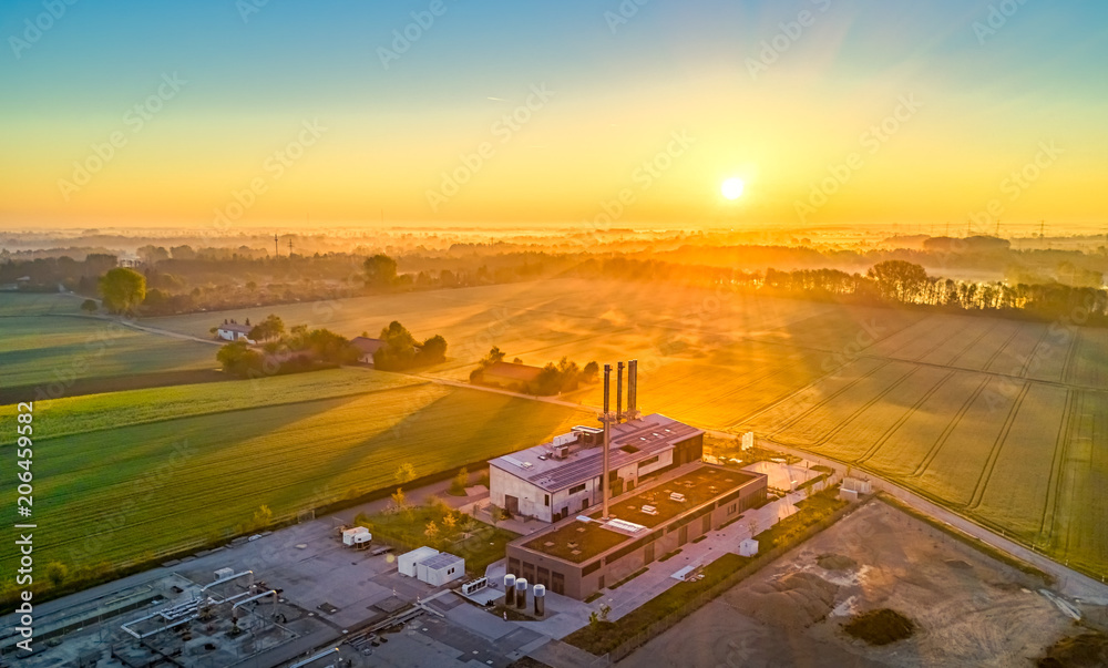 Fototapeta premium Zur goldenen Stunde am frühen Morgen, eine Fabrik vor einem Feld im Hintergrund bei gelb glühender Sonne vor einem See von oben als Luftaufnahme einer Drohne