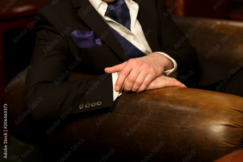a man in a black suit is sitting in a chair. hands folded on the back of the chair. hands of a man in a suit on a chair. style