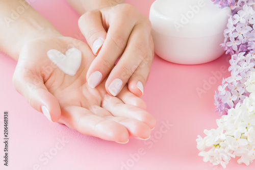 Young, perfect, clean woman's hands. White jar of natural herbal skin cream on pastel pink background. Heart shape created from cream. Love a body. Beautiful branches of lilac blossoms. Fresh flowers.