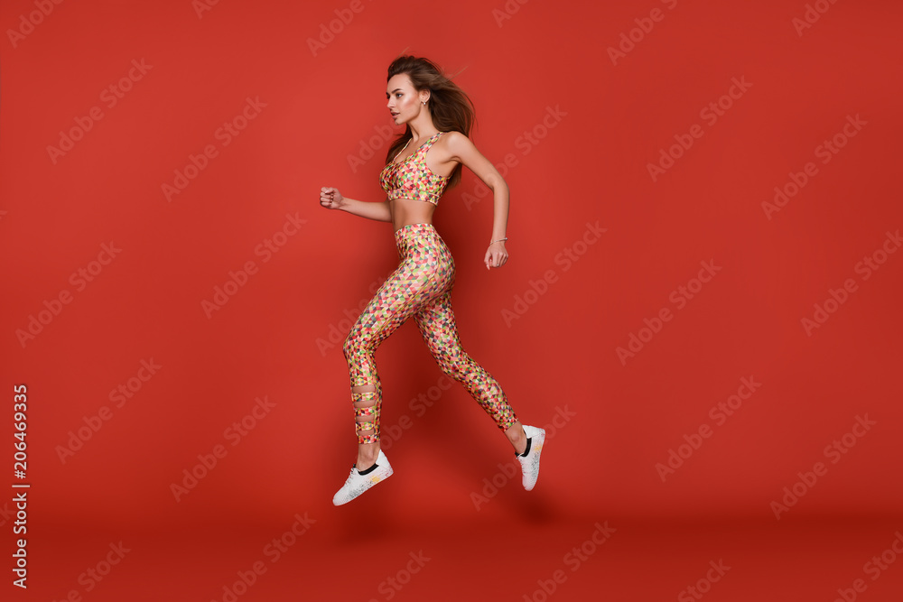 Slim sporty young woman in sport outfit, tight colorful leggings and top with geometrical patterns in a motion, running, jumping high, isolated in studio, bright red background