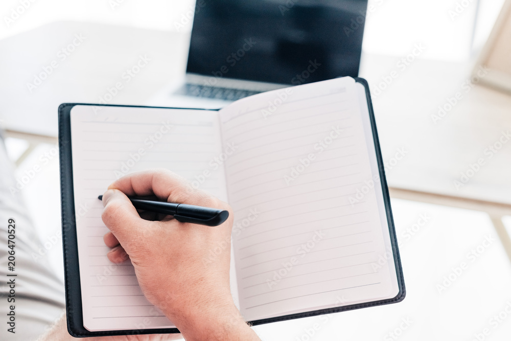 Fototapeta premium cropped image of man writing in textbook at table with laptop