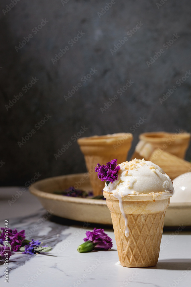 Homemade vanilla ice cream in small waffle cup served with purple edible flowers and metal spoon in ceramic plate on white marble kitchen table.