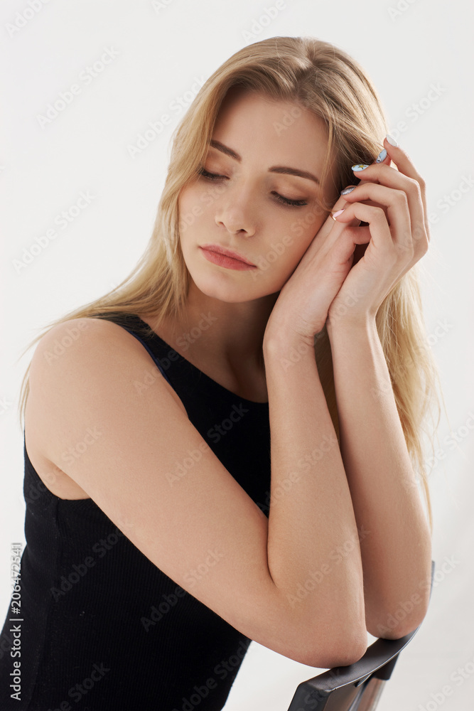 Young beautiful sexy woman in blue jeans shorts and black shirt posing and sitting on simple black chait in white studio. Fashion model with long straight hair and closed eyes. Young beautiful sexy woman in blue jeans shorts and black shirt posing and sitting on simple black chait in white studio. Fashion model with long straight hair and closed eyes.