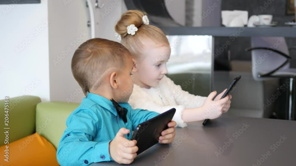 Cute kids boy and girl watch use tablet computer display sitting at cafe table Cute kids boy and girl watch use tablet computer display sitting at cafe table