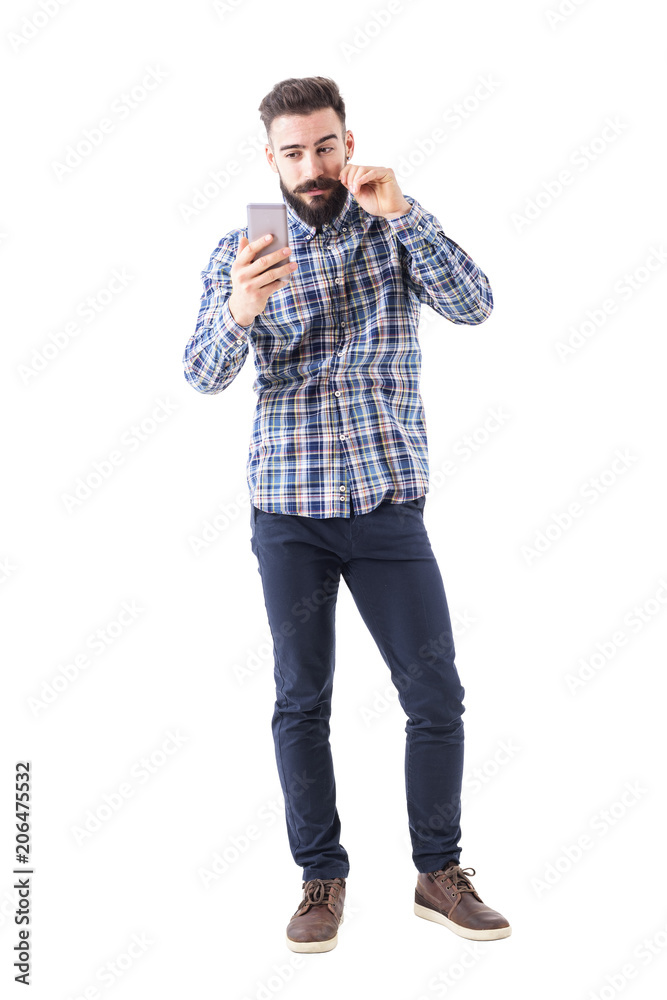 Bearded hipster man twirling mustache using mobile phone reflection as mirror. Full body isolated on white background. Bearded hipster man twirling mustache using mobile phone reflection as mirror. Full body isolated on white background.