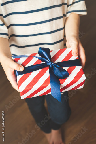 Close up view of boy holding gift box wrapped in stripes paper and tied with blue bow. Boy giving a gift.