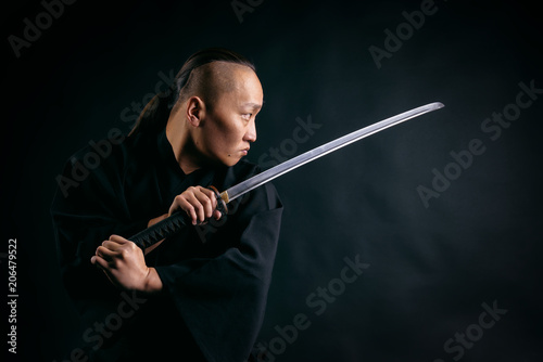 Asian man with a sword in his hands in a black robe against a black background