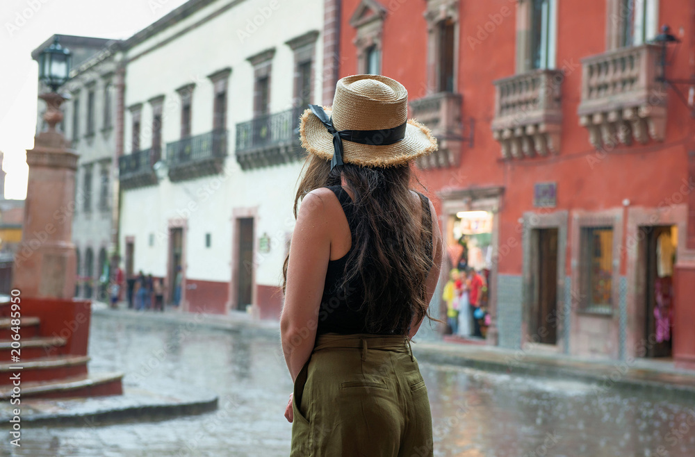 young woman in hat looking out onto street at town square in San Miguel de Allende, Mexico, during rain, with colonial buildings in background