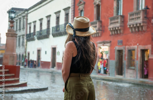 young woman in hat looking out onto street at town square in San Miguel de Allende, Mexico, during rain, with colonial buildings in background