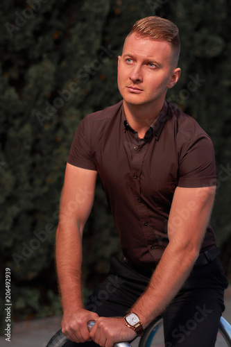 Young and serious businessman wearing T-shirt and black trousers sitting on the street and thinking. Men's fashion.