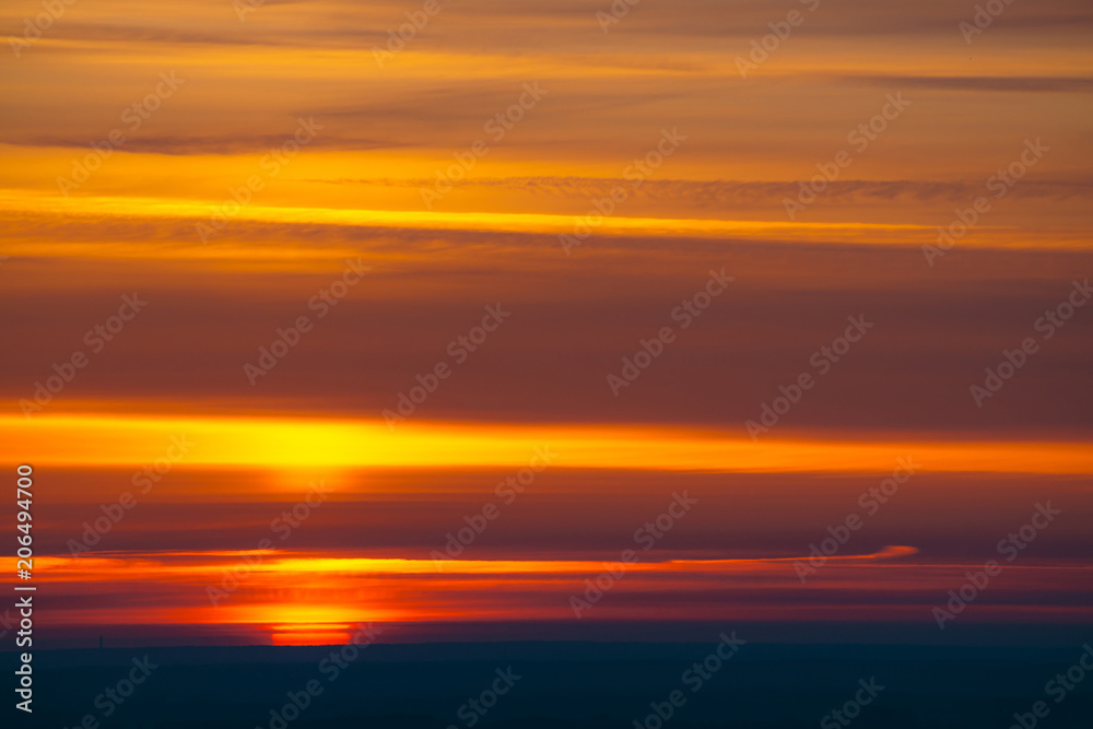 Big red sun circle rises out from behind dark horizon on background varicolored clouds of warmly shades. Beautiful background of dawn on picturesque cloud sky.