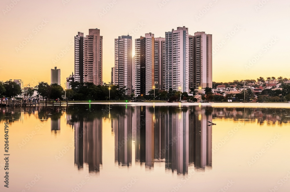 Growing city with few tall buildings reflected on the water of a lake, sunset hour - golden hour. Campo Grande MS, Brasil