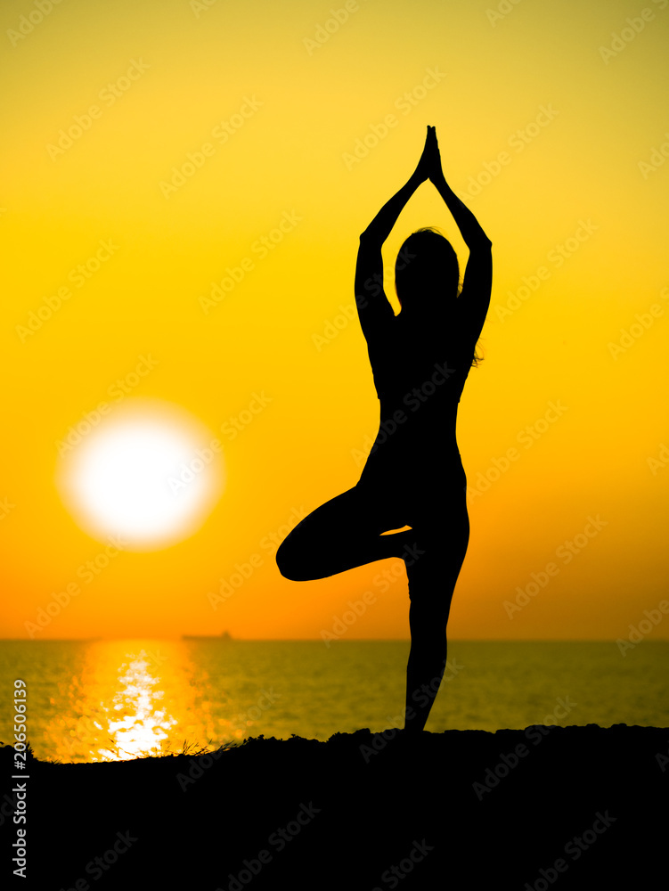 Young slim girl practicing yoga on mountain against ocean or sea at sunrise time. Silhouette of woman in yellow rays of awesome sunset.