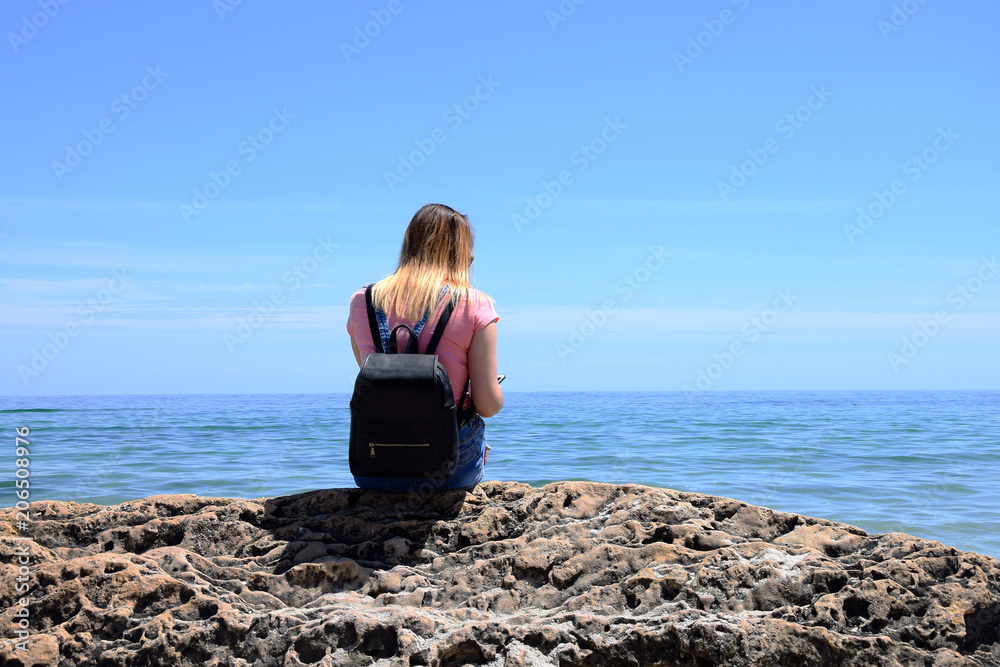  A young girl is sitting with her backpack on a large stone by the sea.