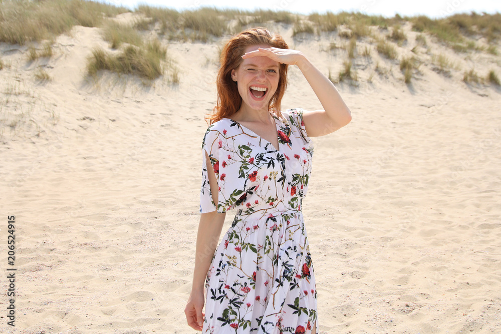 Hübsche rothaarige Frau in einem Kleid steht an einem Strand lächelt zwinkernd und shcützt ihre Augen gegen das Sonnenlicht Hübsche rothaarige Frau in einem Kleid steht an einem Strand lächelt zwinkernd und shcützt ihre Augen gegen das Sonnenlicht
