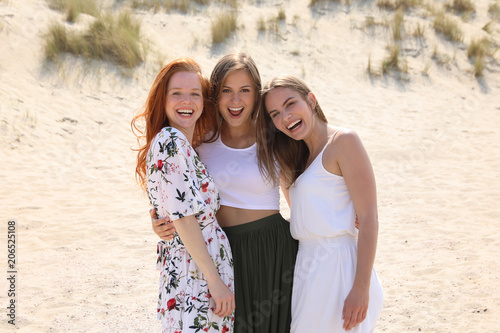 Drei hübsche junge Frauen stehen ARm in Arm an einem Strand und lachen