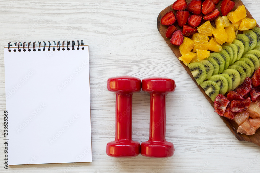 Fitness concept, diet plan. Dumbbells , notebok, chopped fruits on white wooden background, top view. Fitness concept, diet plan. Dumbbells , notebok, chopped fruits on white wooden background, top view.