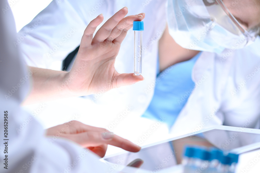 Closeup of scientific research team with clear solution in laboratory. Blonde female chemist holds test tube of glass while her colleague checks results with tablet pc. Blood test, medicine or Closeup of scientific research team with clear solution in laboratory. Blonde female chemist holds test tube of glass while her colleague checks results with tablet pc. Blood test, medicine or