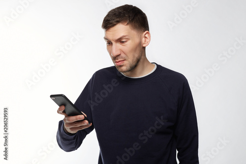Angry irritated young man reads text message on smart phone remind him about bills, needs to pay debts immediately, expresses big rage and displeasure, blows steam,  frowns face in dissatisfaction. © Yulia