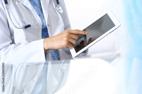Woman doctor using tablet computer while sitting. Reflecting glass table is a working place of physician. Healthcare, insurance and medicine concept