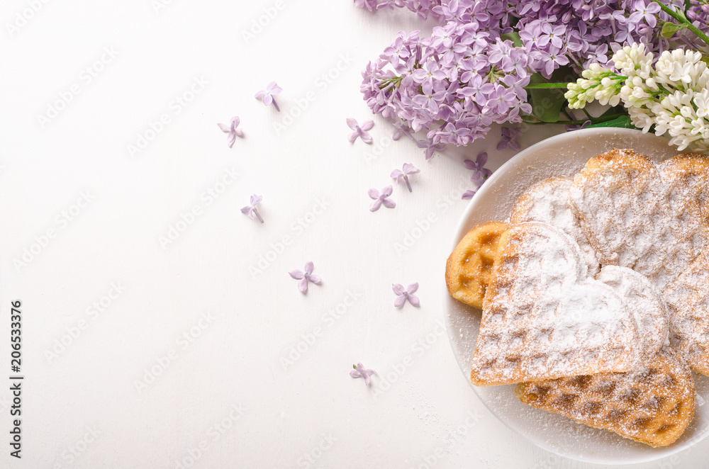 Homemade waffles heart sprinkled with powdered sugar on plate on a white table with flowers. Sweet pastry. Top view Homemade waffles heart sprinkled with powdered sugar on plate on a white table with flowers. Sweet pastry. Top view