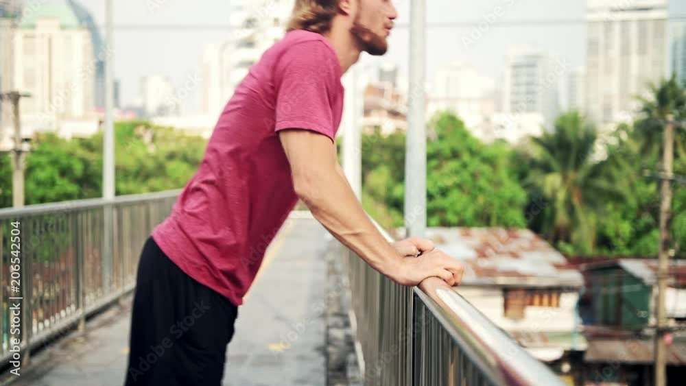 Young LGBT man warming up for a run in urban area. White male in red tshirt stretching arms in a public flyover ready for his run exercise. Natural light. Runner sport concept. Young LGBT man warming up for a run in urban area. White male in red tshirt stretching arms in a public flyover ready for his run exercise. Natural light. Runner sport concept.