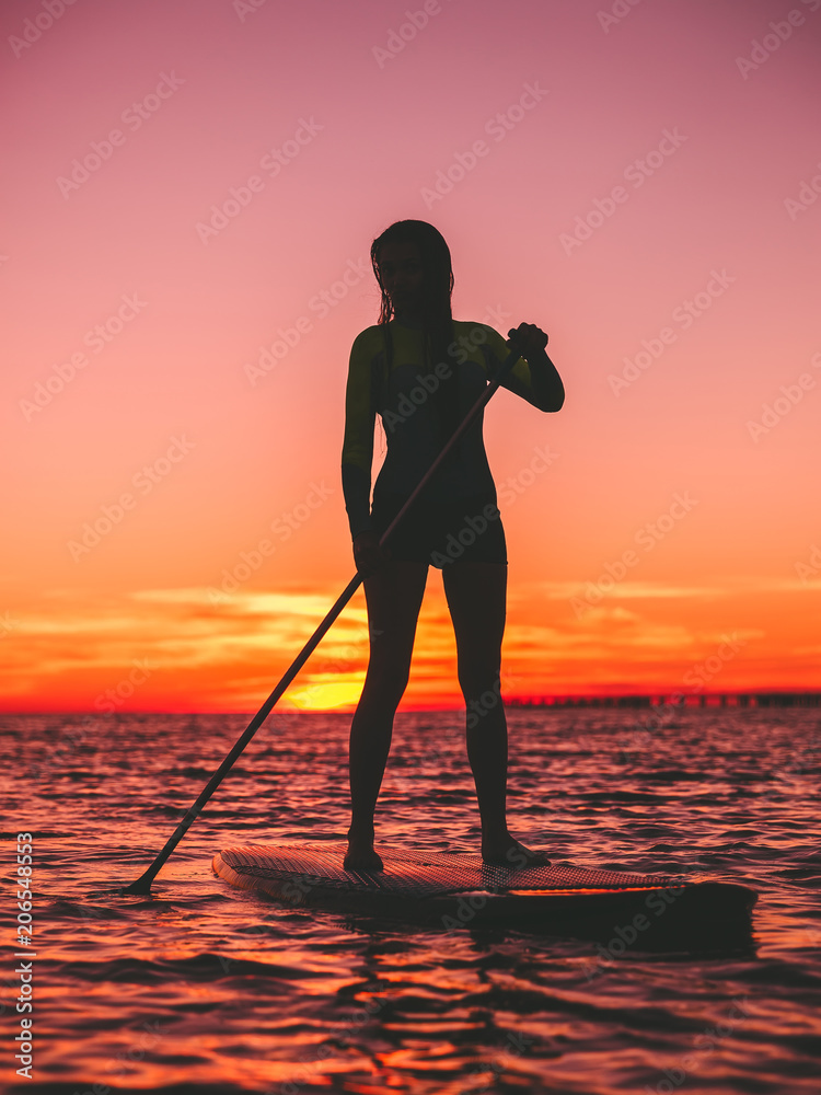 Sporty woman stand up paddle boarding at dusk on a flat warm quiet sea with beautiful sunset